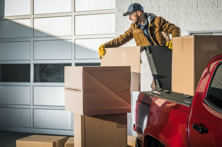 A man packing large cardboard boxes into a red pickup truck in front of a garage door