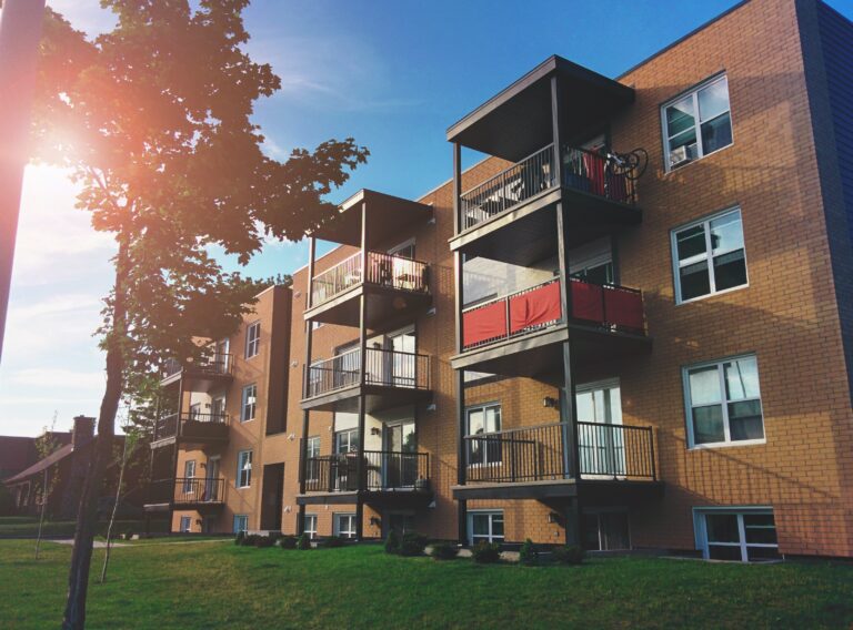 A brown brick 4 storey apartment building with a tree out front on a sunny day